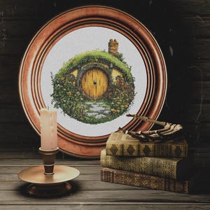 K&ouml;nnte beinhalten: Ein gerahmtes Kreuzstichbild eines Hobbitlochs mit einer gelben T&uuml;r, umgeben von gr&uuml;nem Laub und einem Steinweg. Der Rahmen ist aus braunem Holz und das Bild steht auf einem Holztisch mit einer Kerze und B&uuml;chern.