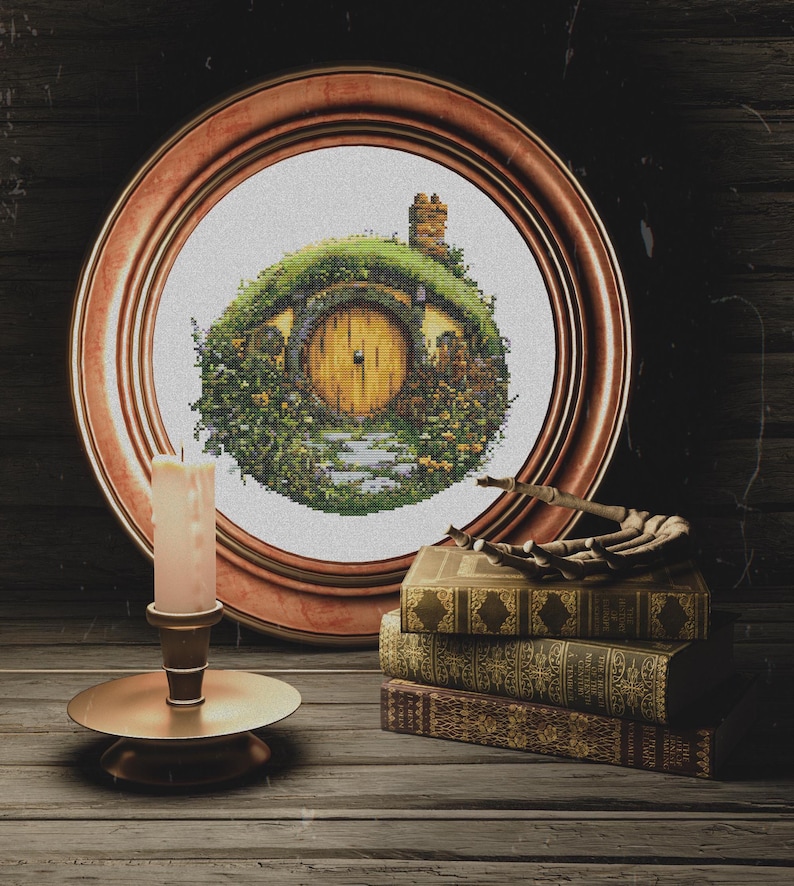 K&ouml;nnte beinhalten: Ein gerahmtes Kreuzstichbild eines Hobbitlochs mit einer gelben T&uuml;r, umgeben von gr&uuml;nem Laub und einem Steinweg. Der Rahmen ist aus braunem Holz und das Bild steht auf einem Holztisch mit einer Kerze und B&uuml;chern.