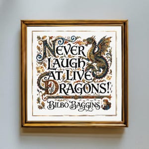 May include: A square cross-stitch embroidery in a gold frame featuring the text "NEVER LAUGH AT LIVE DRAGONS!" and "BILBO BAGGINS". The design includes a detailed, multi-colored dragon breathing fire, intricate floral scrollwork, and a small figure of a character.