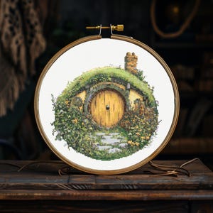 K&ouml;nnte beinhalten: Ein Kreuzstich-Stickrahmen mit einer Darstellung eines Hobbitlochs, einem runden, gr&uuml;n bewachsenen Haus mit einer runden Holzt&uuml;r. Das Haus ist von gr&uuml;nen Pflanzen und Blumen umgeben.