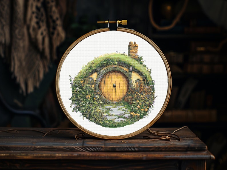 K&ouml;nnte beinhalten: Ein Kreuzstich-Stickrahmen mit einer Darstellung eines Hobbitlochs, einem runden, gr&uuml;n bewachsenen Haus mit einer runden Holzt&uuml;r. Das Haus ist von gr&uuml;nen Pflanzen und Blumen umgeben.