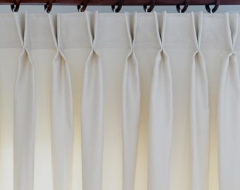 May include: Cream-colored, pleated curtains hanging from a dark brown wooden rod with matching rings. The curtains are neatly folded and create vertical lines, with soft shadows. The fabric appears to be a light, opaque material.