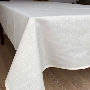 Puede incluir: Mantel de mesa blanco crema con una textura sutil y un borde blanco. El mantel está drapeado sobre una mesa de madera.
