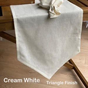 Peut inclure: Un chemin de table en lin blanc crème avec une finition triangulaire. Le chemin de table est posé sur une table en bois avec une serviette blanche pliée en forme de triangle sur le dessus.