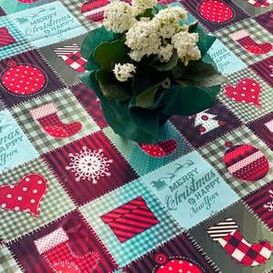Op de afbeelding: Een vierkant kersttafelkleed met een patchwork ontwerp. De vierkanten tonen vakantiemotieven zoals kousen, ornamenten en harten in rood, wit en blauwgroen. Een potplant met witte bloemen staat op het tafelkleed. De tekst "Merry Christmas Happy New Year" is zichtbaar.