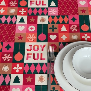 Könnte beinhalten: Ein festlicher Weihnachtstischläufer mit einem geometrischen Muster aus Quadraten und Rauten in Rot, Rosa, Grün und Beige. Das Wort "JOYFUL" ist in großen Buchstaben dargestellt. Weiße Teller und eine Gabel sind auf dem Tischläufer platziert.