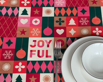May include: A festive Christmas tablecloth with a geometric pattern of squares and diamonds in red, pink, green, and beige. The word "JOYFUL" is displayed in large letters. White plates and a fork are set on the tablecloth.