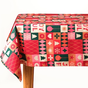 May include: A festive Christmas tablecloth with a repeating pattern of geometric shapes in red, green, pink, and beige. The design includes Christmas ornaments, snowflakes, and the word "JOYFUL". The tablecloth is draped over a wooden table.