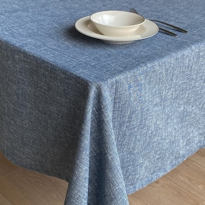 Op de afbeelding: Een blauwe linnen tafelkleed met een witte kom en een wit bord erop. Het tafelkleed heeft een subtiel textuurpatroon.