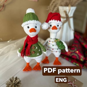Op de afbeelding: Twee handgemaakte gehaakte ganzenbeeldjes, één met een groen-witte hoed, een rode sjaal en een groene jas met knopen. De andere gans draagt een rode kerstmuts en is versierd met lichtslingers. De tekst "PDF pattern ENG" is zichtbaar.