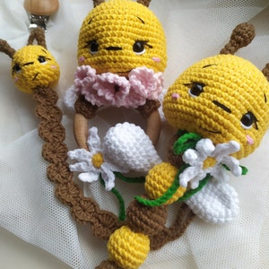 May include: A crocheted bee baby toy with a wooden ring, a daisy flower, and a brown and yellow beaded chain. The bee has a yellow body, brown antennae, and a pink flower on its head.