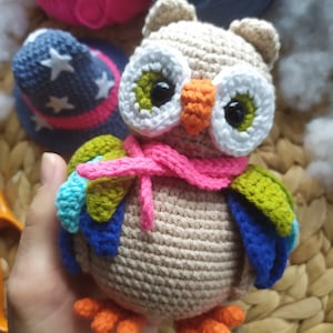 May include: A crocheted owl toy with a pink scarf, green and blue wings, and orange feet. The owl has large white eyes with black pupils and a bright orange beak.