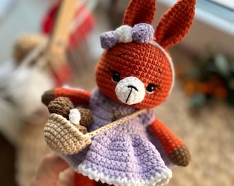 Puede incluir: Una ardilla de crochet naranja que lleva un vestido morado con ribete blanco y una cesta marrón con bolas marrones y blancas. La ardilla tiene un lazo blanco con una flor morada en la cabeza.