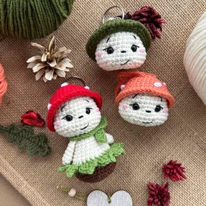 May include: Crocheted mushroom keychains in red, orange, and green hats with smiling faces. One mushroom has a green scarf and a brown base. The keychains are on a burlap surface with yarn and decorative elements.