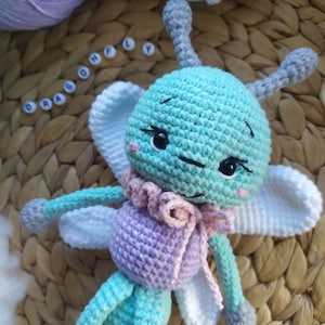 Può includere: Un giocattolo di libellula all'uncinetto realizzato con filato di colore azzurro, viola e bianco. La libellula ha ali grandi, un corpo lungo e una piccola testa con occhi neri. Il giocattolo è seduto su una superficie di vimini intrecciato.