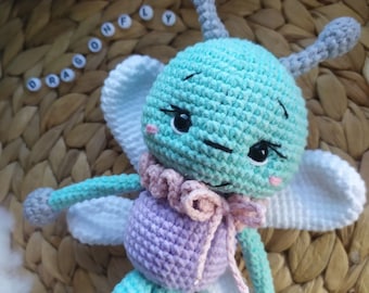 May include: A crocheted dragonfly toy with light blue, purple, and white yarn. The dragonfly has large wings, a long body, and a small head with black eyes. The toy is sitting on a woven wicker surface.