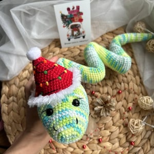 May include: A crocheted snake toy in green, blue, and yellow, wearing a red Santa hat with white trim and a white bobble. The snake has black eyes and is on a woven surface. A Christmas card is in the background.