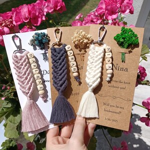 Könnte beinhalten: Drei Brautjungfern-Anfragekarten mit einem rosa, weißen und grauen Quasten-Schlüsselanhänger mit Holzperlen, die die Namen "Darlene", "Sarah" und "Nina" buchstabieren. Die Karten sind braun mit weißem Text und beinhalten die Frage "Willst du meine Brautjungfer sein?" und das Datum "3/29/2025".