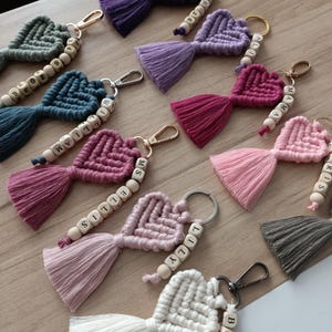 Pode incluir: Uma coleção de porta-chaves de macramé em forma de coração em várias cores, incluindo roxo, lavanda, magenta, rosa, verde sálvia, azul e branco. Cada porta-chaves apresenta um coração atado, uma borla a condizer e contas de madeira com letras ou números.