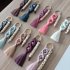 Peut inclure: Porte-clés en macramé avec des perles en bois épelant des noms et des dates, avec des perles en forme d'œil. Chaque porte-clés a un pompon de différentes couleurs, dont le rose, le violet et le bleu. Les porte-clés sont attachés à des anneaux dorés.