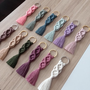 Könnte beinhalten: Eine Sammlung von Makramee-Schlüsselanhängern in verschiedenen Farben, darunter Rosa, Grün, Blau, Lila und Weiß. Jeder Schlüsselanhänger hat ein gewebtes Design und einen passenden Quastenanhänger. Die Schlüsselanhänger sind auf einer hellen Holzoberfläche angeordnet.