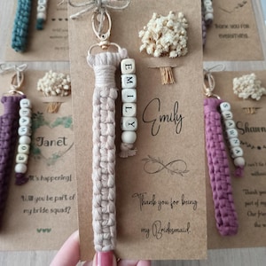 May include: A beige macrame keychain with a gold-tone lobster clasp and a side strand of white square beads spelling the name EMILY. The keychain is attached to a rectangular kraft paper card featuring the name Emily and the text Thank you for being my Bridesmaid.