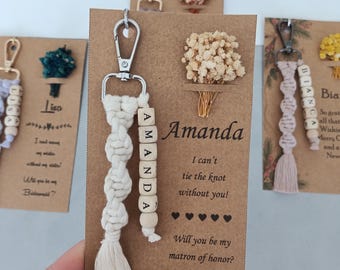 May include: A macrame keychain with a silver lobster clasp, cream-colored cord, and wooden letter beads spelling AMANDA, attached to a brown card featuring a dried flower bouquet and the text Will you be my matron of honor?