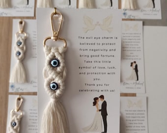 May include: A macrame keychain featuring two blue and white evil eye beads, a gold-tone lobster clasp, and a cream-colored tassel. It is attached to a white card printed with two doves, a wedding illustration, and the text Dion + Michael February 3, 2026.