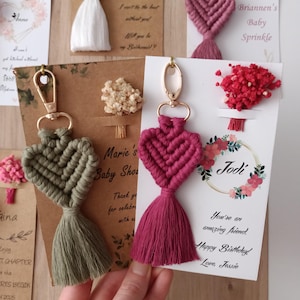 Könnte beinhalten: Makramee-Herz-Schlüsselanhänger in Rosa, Grün und Weiß mit goldfarbenen Verschlüssen. Jeder Schlüsselanhänger hat eine Quaste und ist mit einer personalisierten Karte versehen. Die Karten haben florale Akzente und handschriftlichen Text.