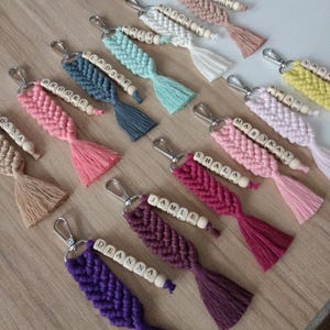 May include: A collection of handmade macrame keychains in various colors, including pink, blue, purple, and white. Each keychain features a braided design, wooden beads with names, and a tassel. The keychains are attached to silver-colored metal clasps.