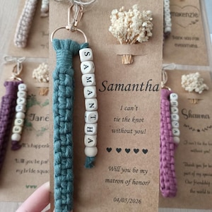 May include: A teal macrame keychain with a gold-tone clasp and a strand of white letter beads spelling SAMANTHA, attached to a brown card featuring dried baby's breath and the text I can't tie the knot without you! Will you be my matron of honor?