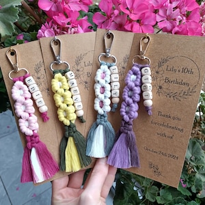 May include: Four colorful keychains with names spelled out in wooden beads. Each keychain has a tassel and a flower design. The card reads "Lily's 10th Birthday" and "Thank you for celebrating with us! October 24th 2024".
