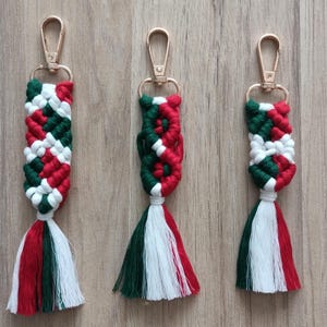 Pode incluir: Três porta-chaves de macramé com um padrão entrançado em cordão vermelho, verde escuro e branco. Cada porta-chaves está preso a um mosquetão de metal dourado e termina na parte inferior com uma borla multicolorida em vermelho, verde e branco.