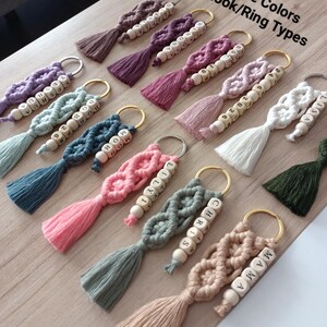 Può includere: Una collezione di portachiavi in macramè in vari colori, ognuno con un nappa e perline di legno con lettere. I portachiavi sono esposti su una superficie di legno chiaro. Il testo sull'immagine recita "43 Colori Corda 10 Tipi di Gancio/Anello."