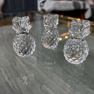 May include: Three clear glass pineapple-shaped candle holders. The candle holders have a textured, diamond-patterned body and a detailed leafy top. They are arranged on a reflective surface, creating a play of light and shadow.