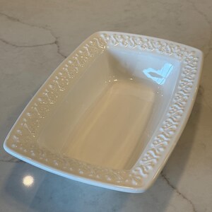 Peut inclure: Un plat de service rectangulaire en céramique blanc cassé présentant un motif floral en relief complexe sur tout le bord. L'intérieur du plat est lisse et brillant, avec un bassin profond et des côtés légèrement évasés.