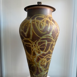 Puede incluir: Un jarrón alto de cerámica marrón con tapa. El jarrón tiene una superficie texturizada con diseños abstractos amarillos y verdes. La tapa tiene un asa pequeña. El jarrón se exhibe sobre un pedestal blanco.