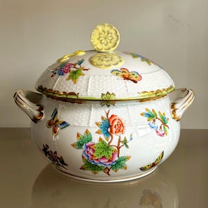 以下が含まれることがあります： カラフルな花柄と蝶の模様が施された、蓋付きの白い磁器製のサービングボウル。ボウルは金色のトリムとハンドルを備え、織りかごのような質感です。蓋には黄色の花のノブが付いています。