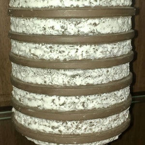 May include: A cylindrical ceramic vase with a textured white surface and horizontal brown bands. The vase has a wide, rounded base and a slightly narrower top. The overall design is simple and modern, with a focus on texture and form.