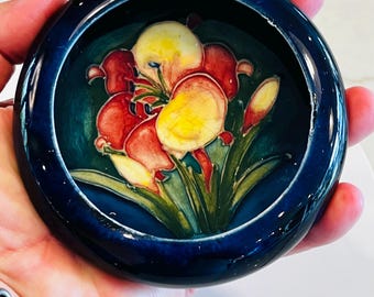 May include: A small, round, dark blue ceramic dish with a floral design. The interior features a painting of red and yellow flowers with green leaves against a dark teal background. The dish is held in a person's hand.