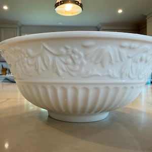 Peut inclure: Un grand bol en céramique blanche présentant un motif en relief de feuilles et de baies sur le bord supérieur, avec une série de cannelures verticales décorant la partie inférieure de l'extérieur, créant un aspect texturé et classique.