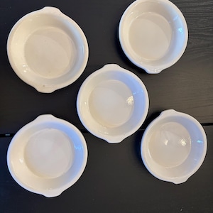 以下が含まれることがあります： 小さな丸い白いセラミック製の皿が5つ。縁が盛り上がり、小さな取っ手が2つ付いています。暗い面に配置されています。これらの皿は、個々の料理を盛り付けるのに適しています。