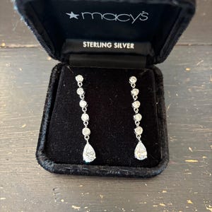 May include: A pair of sterling silver drop earrings displayed in a black velvet Macy's jewelry box. Each earring features a series of round, clear stones leading to a pear-shaped stone at the bottom. The box reads "STERLING SILVER".