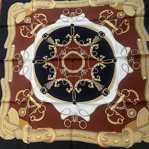 May include: Square silk scarf with an equestrian design. The scarf features a repeating pattern of gold-coloured horse bits, chains, and belts against a black, brown, white, and gold background. The scarf has a decorative border.
