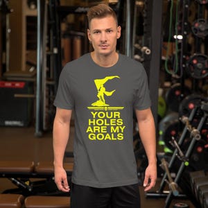 Könnte beinhalten: Ein dunkelgraues T-Shirt mit einer gelben Grafik einer Person, die eine Flagge mit dem Text "YOUR HOLES ARE MY GOALS" hält.