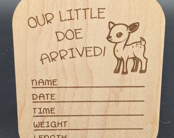 Puede incluir: Un letrero de madera con el texto "Our Little Doe Arrived!" y una ilustración de un ciervo de dibujos animados. El letrero tiene líneas en blanco para el nombre, la fecha, la hora, el peso y la longitud del bebé.