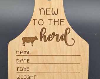 Puede incluir: Una etiqueta de vaca de madera con el texto "New to the herd" y espacios para escribir el nombre, la fecha, la hora, el peso y la longitud.