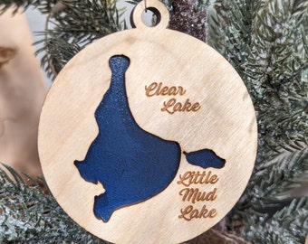 Puede incluir: Un adorno navideño de madera con un contorno azul del lago Clear y el lago Little Mud. El adorno está colgado en un árbol de Navidad.
