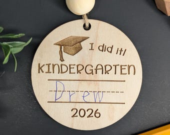 Puede incluir: Adorno circular de madera con el texto "I did it! KINDERGARTEN Drew 2026" y una ilustración de un birrete. El adorno está sujeto a una cuerda de hilo natural con una cuenta de madera. Un lápiz amarillo está en la esquina inferior derecha.