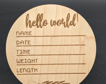 Puede incluir: Una placa redonda de madera con el texto "hello world!" grabado en ella. Hay líneas en blanco para "name", "date", "time", "weight", y "length". Un pequeño diseño de espiga de trigo está grabado en la parte inferior.